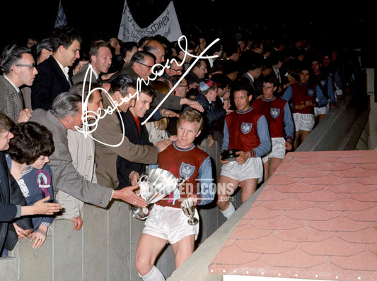 Historic colour photograph of Bobby Moore carrying the FA Cup following West Ham United’s 1964 triumph at Wembley, complete with a reproduced Bobby Moore autograph.
